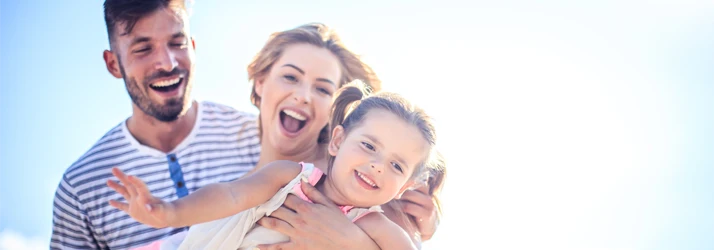 Three adults outdoors holding a young child, standing close together against a bright sky background.